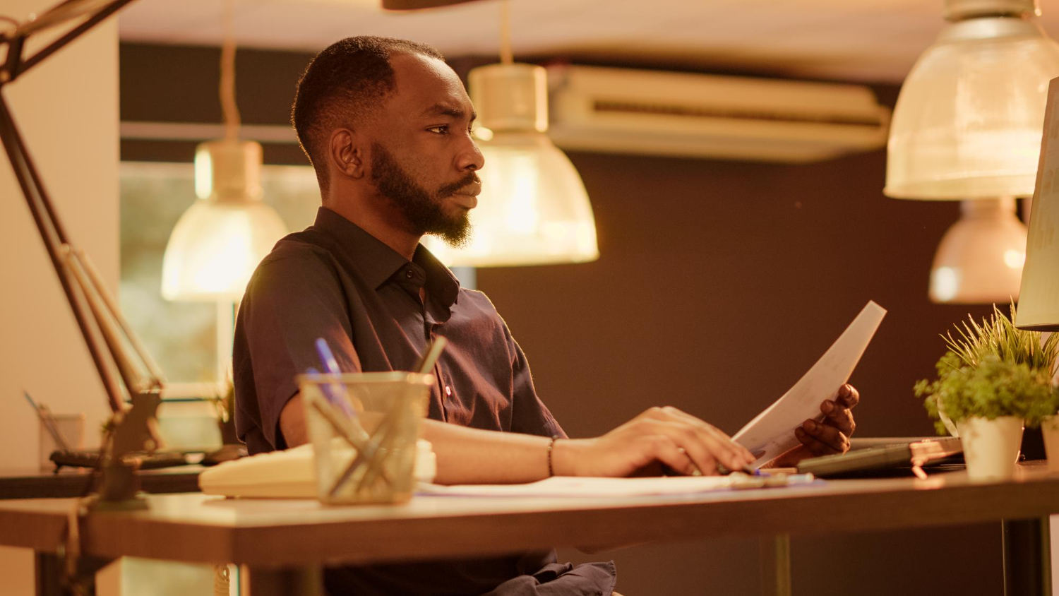 Homem negro de barba bem aparada, vestindo uma camisa social escura, está concentrado em sua mesa de trabalho. Com um olhar atento, ele segura um documento enquanto analisa informações no computador, em um ambiente de escritório iluminado por luminárias modernas. O espaço organizado, com itens como um caderno, lápis e pequenos vasos de plantas, transmite uma atmosfera profissional e produtiva, refletindo um momento de foco e estratégia. A cena representa o conceito de planejamento de carreira, destacando a importância da análise e organização para o crescimento profissional