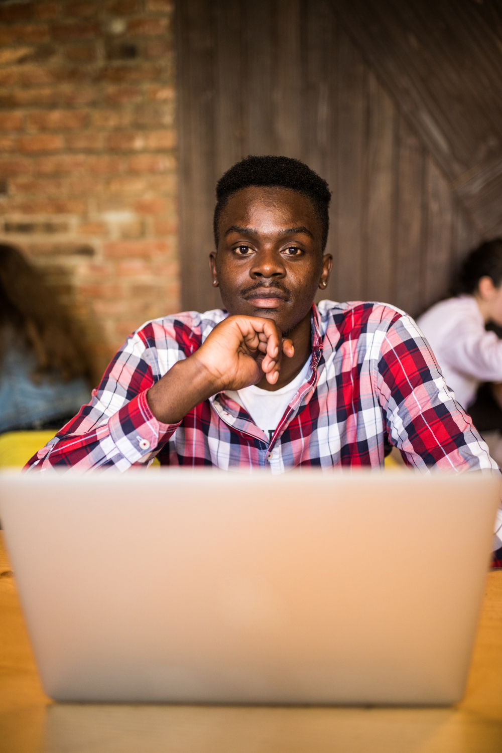 Homem negro usando uma camisa xadrez vermelha e branca, sentado em frente a um laptop em um ambiente casual. Ele está com uma expressão séria e olhar focado, representando a ideia de concentração e produtividade. A imagem reflete a importância de criar conteúdo inclusivo com IA, promovendo a diversidade em ambientes tecnológicos e criativos.