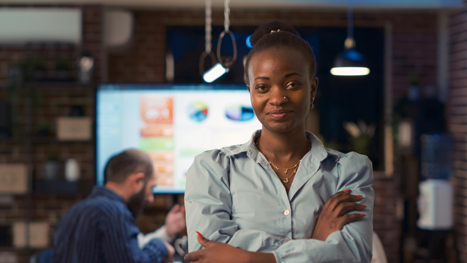 Mulher preta de expressão confiante, vestindo uma camisa azul-clara e com os braços cruzados, se destaca em um ambiente corporativo moderno. Ao fundo, uma tela exibe gráficos e dados de desempenho, enquanto colegas trabalham. A imagem simboliza a atuação de uma líder de marketing generalista, demonstrando estratégia e protagonismo no setor.