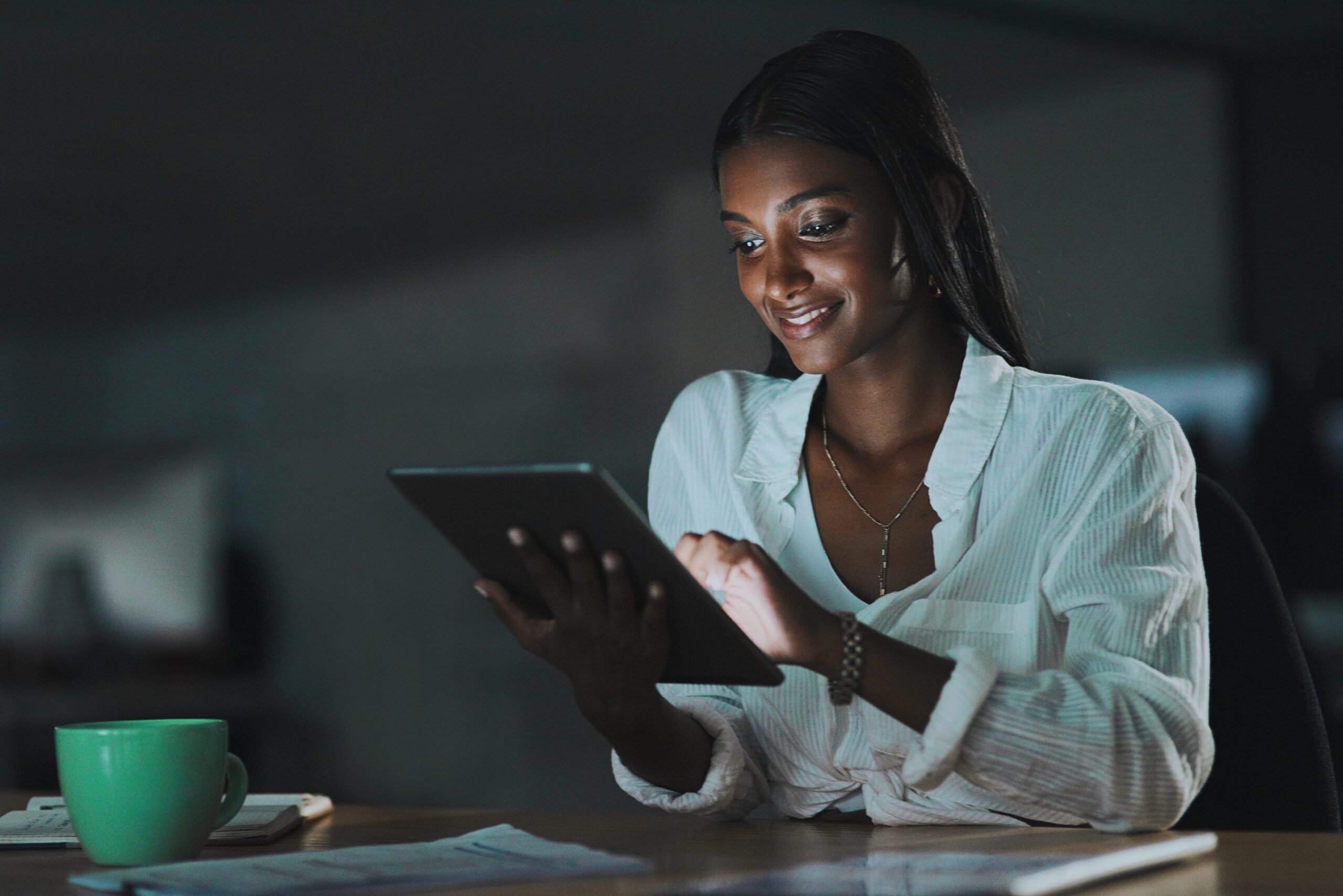 Em um ambiente de trabalho com iluminação baixa, uma mulher negra com cabelos longos e lisos sorri enquanto utiliza um tablet. Ela veste uma camisa branca com amarração na frente e acessórios dourados. A luz da tela ilumina seu rosto, destacando sua expressão concentrada e confiante. Na mesa à sua frente, há uma xícara verde, documentos e um caderno. A cena transmite a força da comunicação quilombola no mundo digital, refletindo como as redes quilombolas utilizam a tecnologia para fortalecer narrativas negras, inovar e manter viva a tradição da resistência.