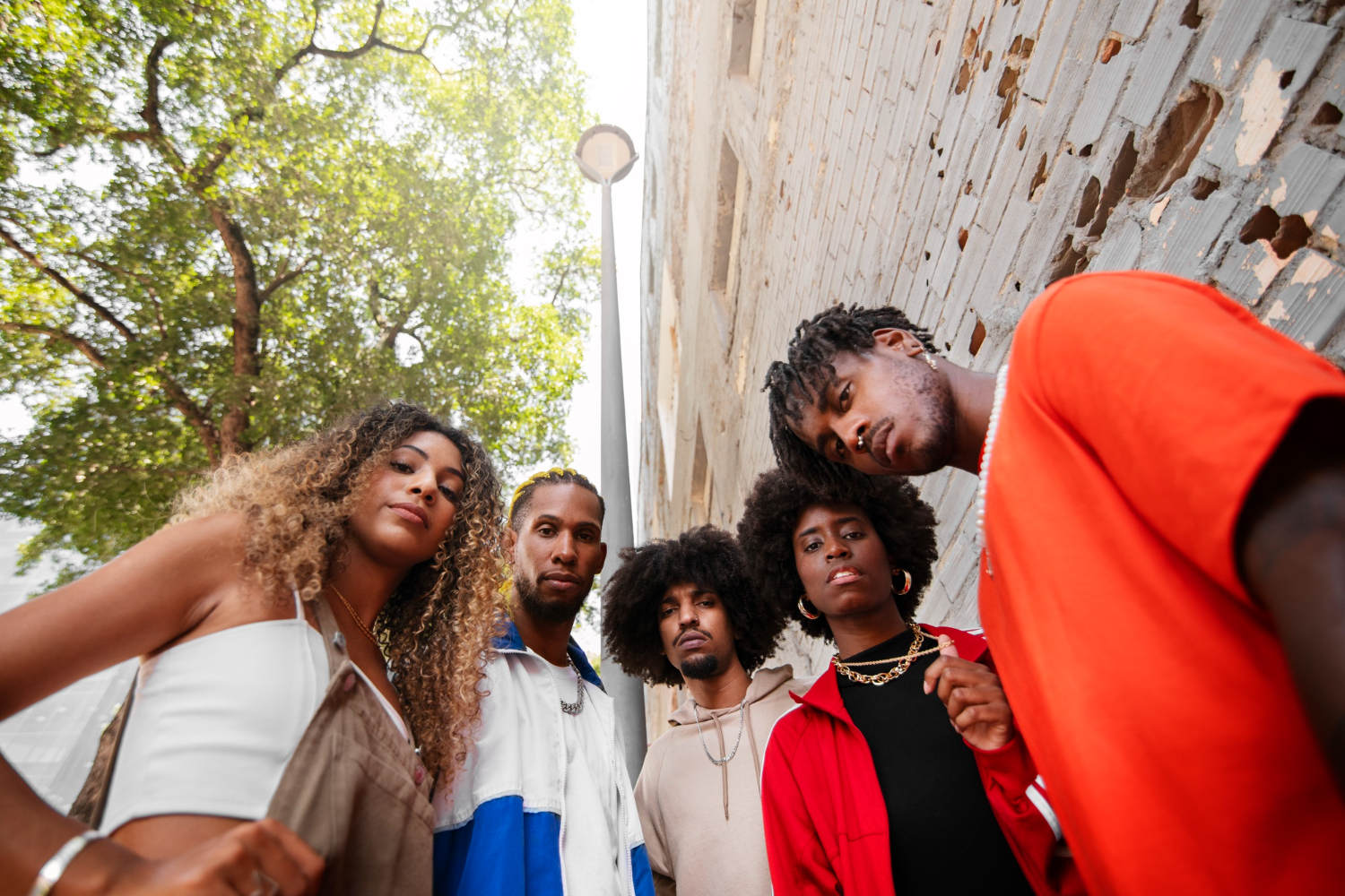 Na imagem, cinco jovens negros posam de forma confiante em um cenário urbano, evidenciando a diversidade e a força da Cultura Negra. Eles vestem roupas modernas e coloridas, com diferentes estilos de cabelo que ressaltam sua identidade. Ao fundo, é possível ver uma parede de tijolos e a copa de uma árvore, compondo um ambiente urbano que realça a criatividade e o orgulho presentes na cultura afro-brasileira.