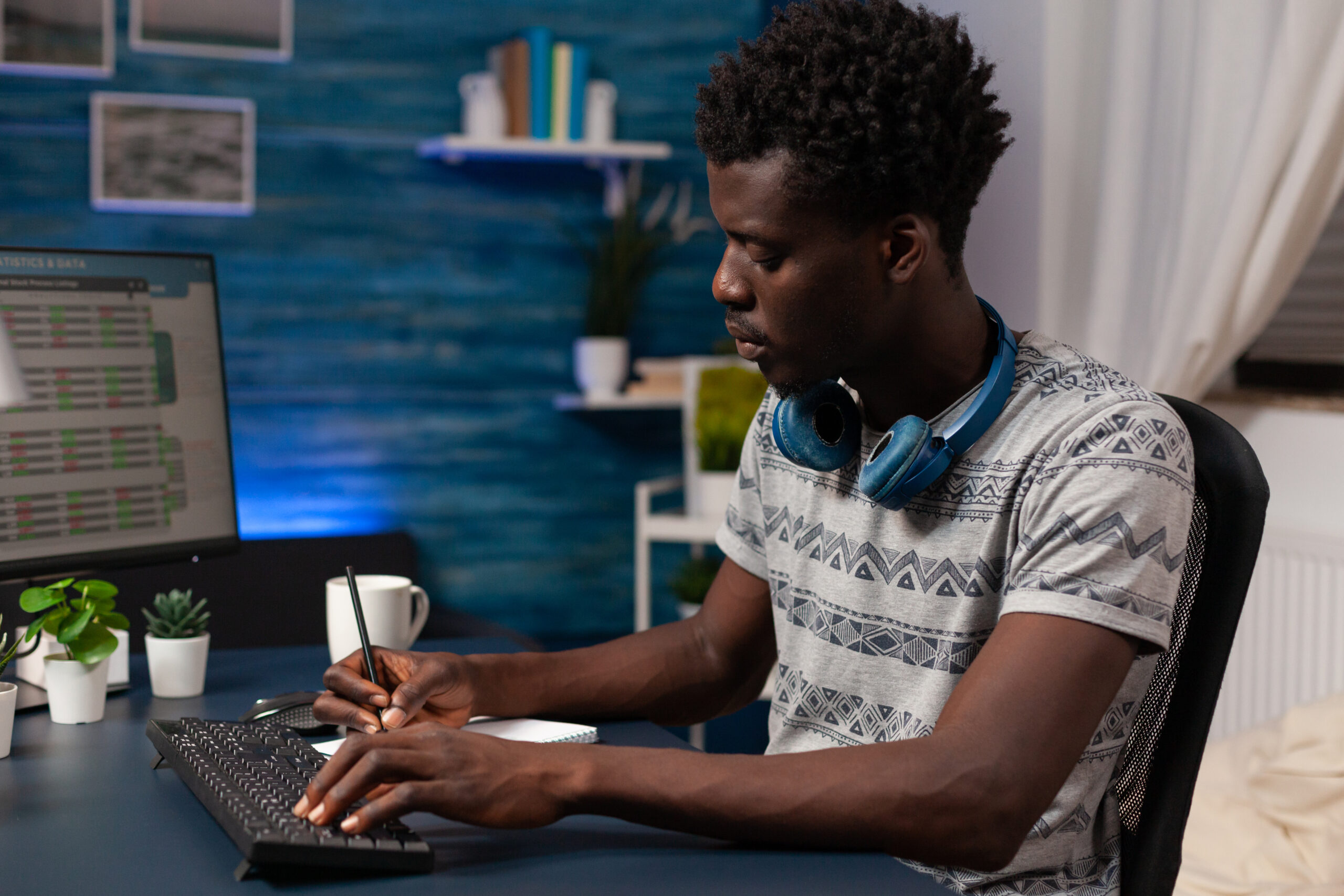Homem jovem preto sentado à mesa, usando fones de ouvido e fazendo anotações em frente ao computador, simbolizando a descentralização da inovação.