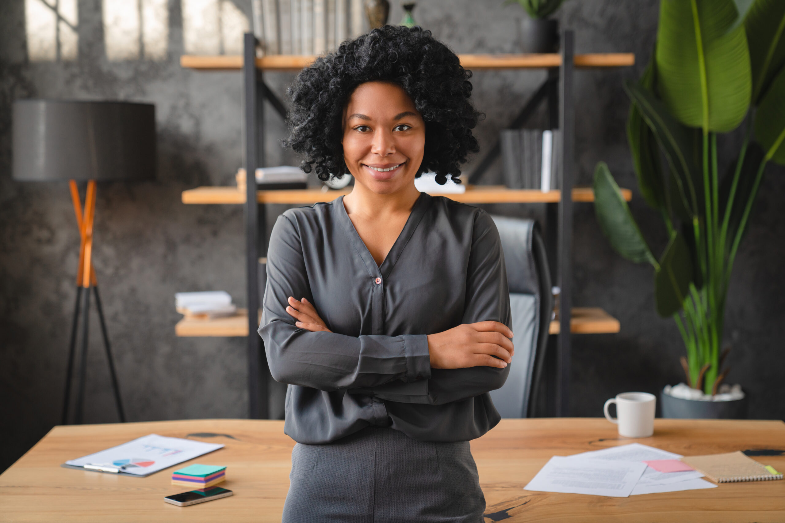 Uma mulher negra, com expressão confiante e postura ereta, está de braços cruzados em frente a uma mesa de escritório, simbolizando a liderança e a **gestão de equipes** em um ambiente corporativo. Ela veste uma camisa social cinza e sorri levemente, transmitindo segurança e profissionalismo. Ao fundo, vê-se uma estante com objetos decorativos e plantas, reforçando a atmosfera moderna e organizada do espaço de trabalho.