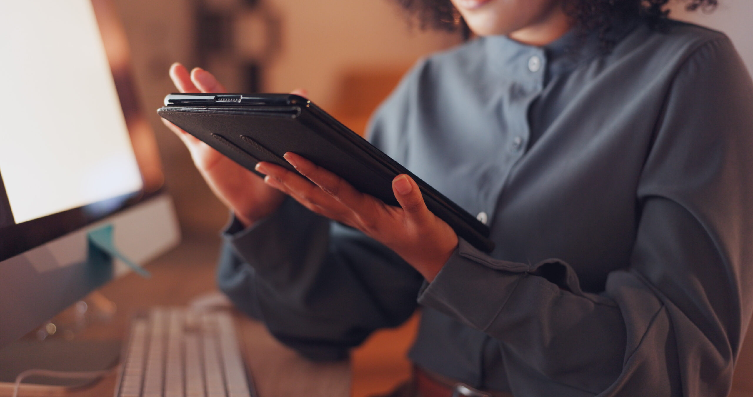Uma mulher preta, vestida com uma camisa cinza, segura um tablet enquanto trabalha em um escritório como social media. Ela está focada em criar e analisar estratégias para redes sociais, demonstrando concentração em suas tarefas diárias.