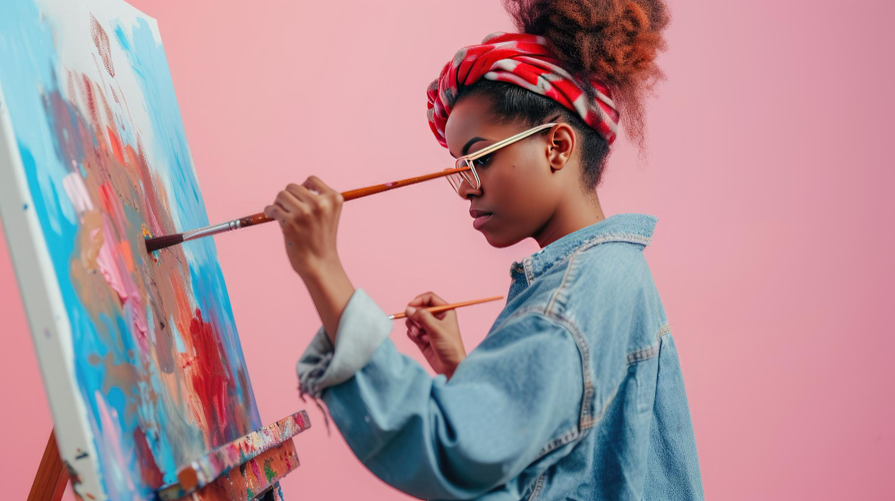 mulher negra pintando um quadro com pincel. O quadro tem cores como azul e vermelho. Ela usa um black em um coque alto e usa um turbante.