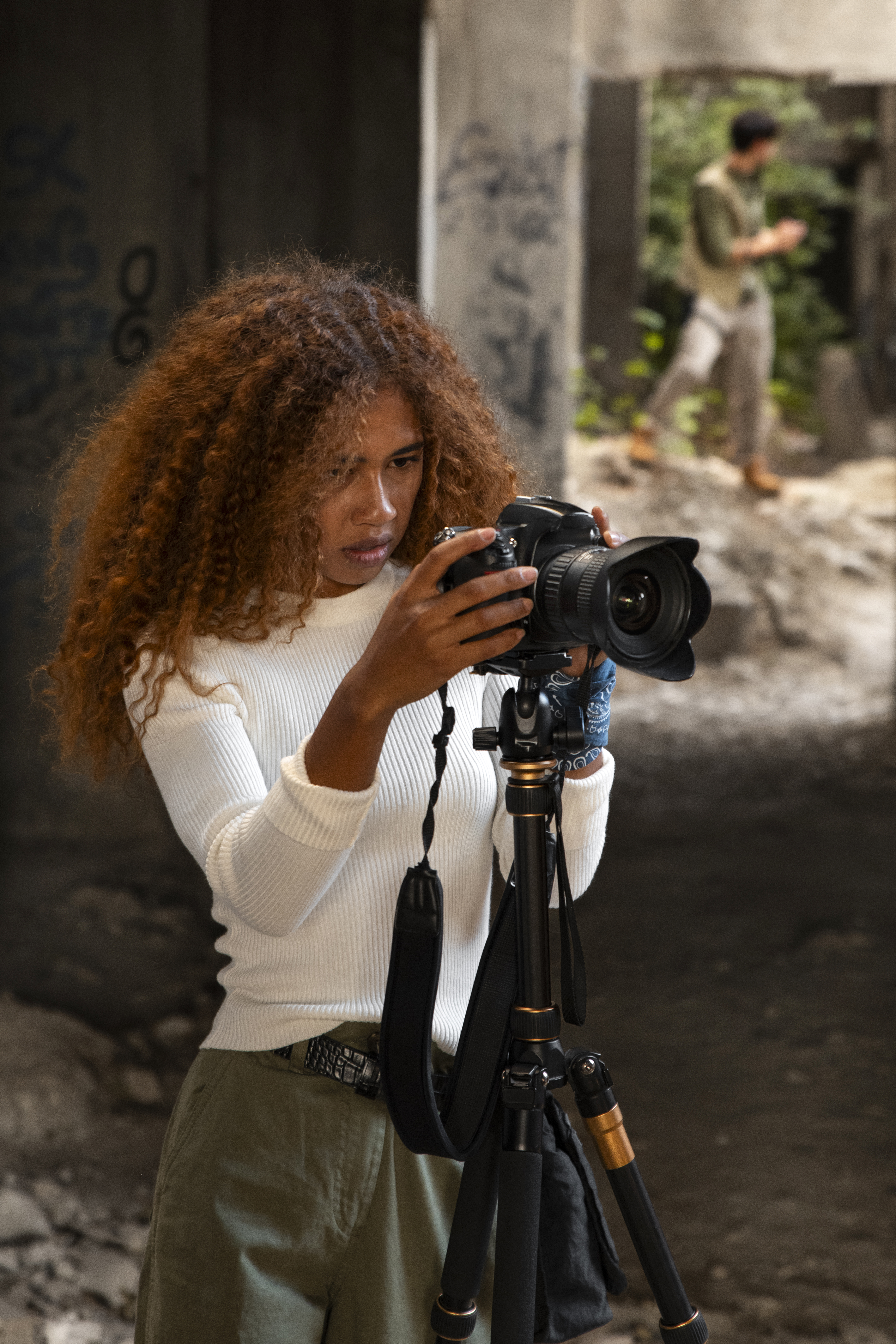 Uma jovem profissional negra do setor audiovisual ajusta sua câmera em um tripé enquanto se concentra na captura da imagem. Com cabelos volumosos e vestindo roupas casuais, ela está em um ambiente urbano e rústico, onde a arte e a técnica do audiovisual se encontram para contar histórias autênticas. Ao fundo, um homem desfocado caminha, acrescentando profundidade à cena. A imagem destaca a representatividade no audiovisual, evidenciando como a diversidade de olhares pode transformar narrativas e carreiras digitais.