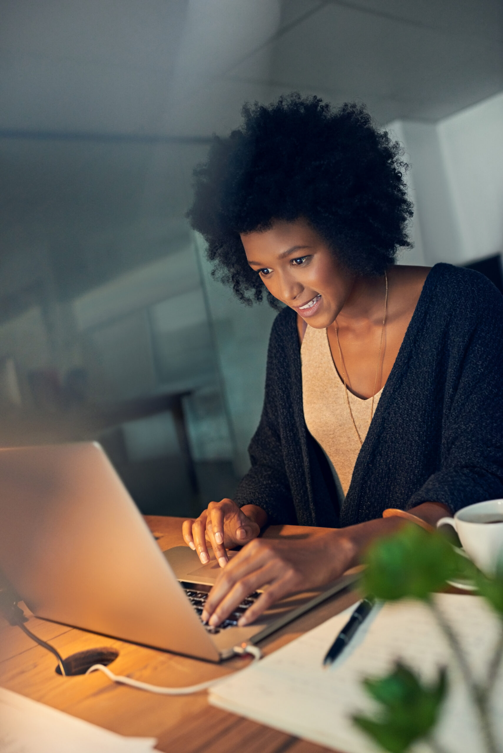 Uma jovem profissional negra, concentrada e sorridente, trabalha em seu laptop em um ambiente de escritório, simbolizando o empoderamento feminino na tecnologia. Papéis, caneta e uma xícara sobre a mesa reforçam seu foco e criatividade, destacando a importância da presença de mulheres em projetos de programação e inovação