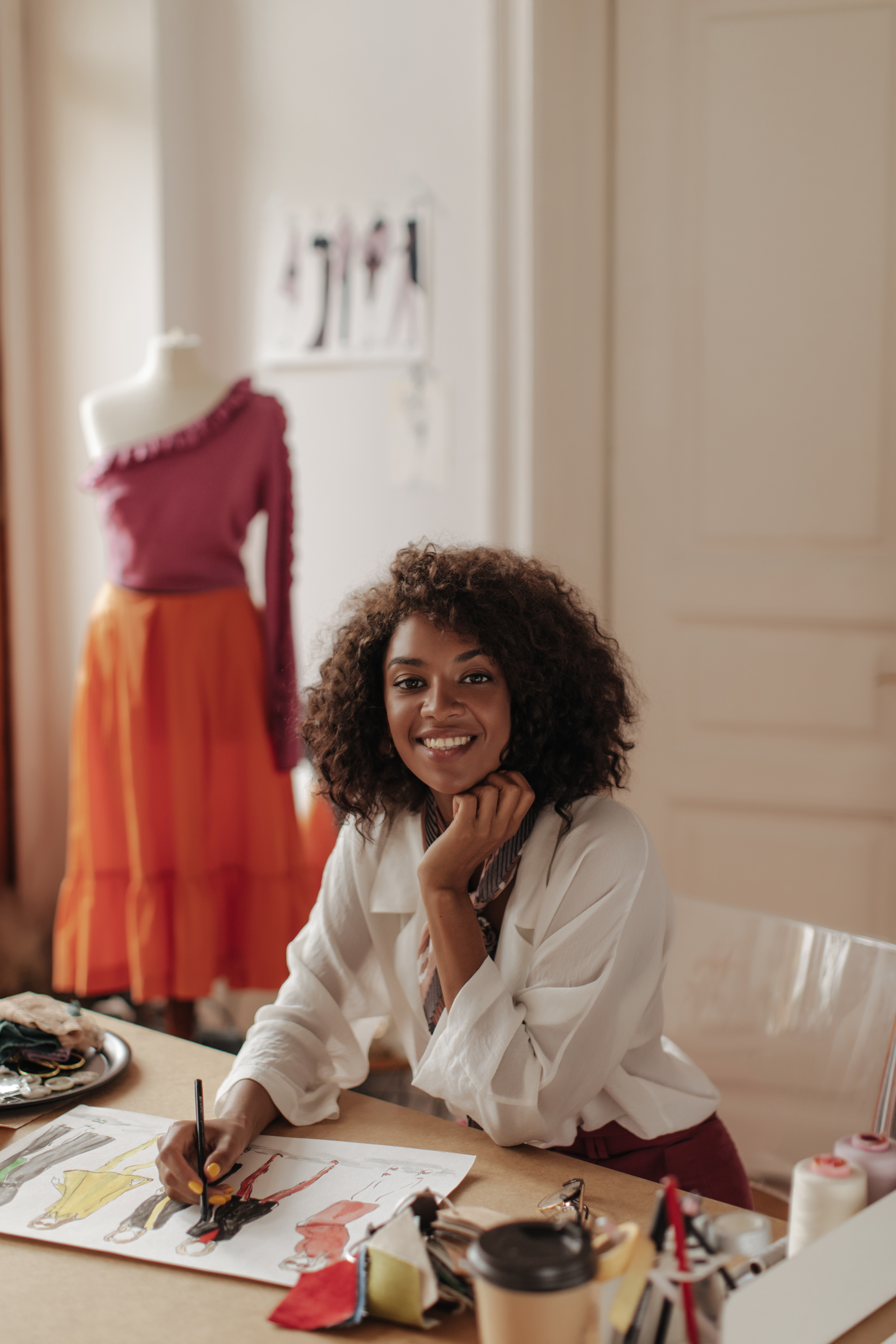 Uma jovem empreendedora negra, sentada em um ateliê de moda, sorri enquanto desenvolve rascunhos coloridos em uma folha de papel. Ao fundo, um manequim exibe roupas vibrantes, evidenciando o potencial do empreendedorismo criativo na criação de novos negócios. Materiais de pintura e costura espalhados pela mesa destacam o processo de inovação e expressão artística no Brasil.