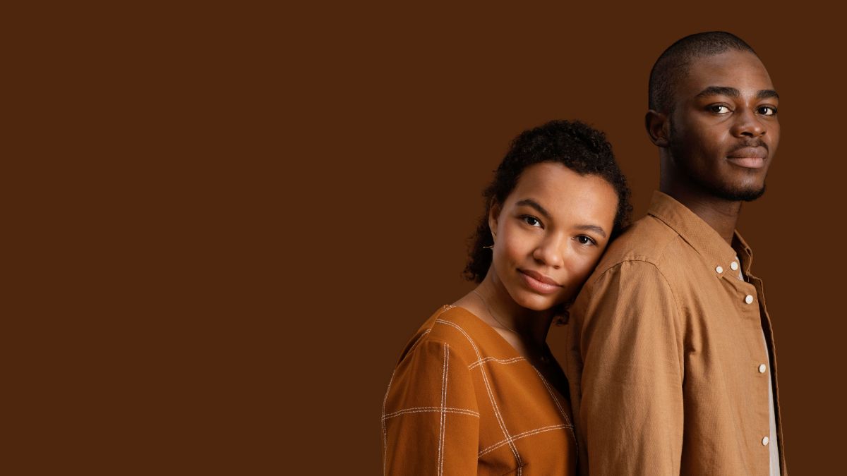 pessoas negras: um homem negro alto vestido de camiseta bege e uma mulher negra um pouco mais baixa que o homem e vestida de camisa marrom, encosta a cabeça em suas costas