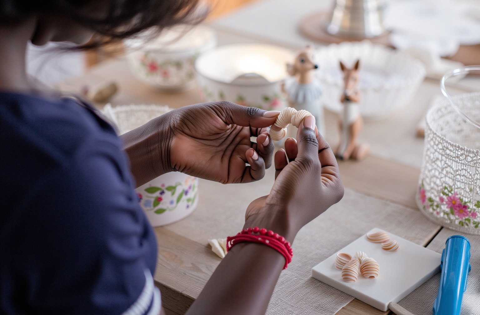 Em uma mesa de trabalho artesanal, vê-se as mãos de uma mulher negra manuseando pequenas peças em formato de conchas, representando a produção artística típica do setor de economia criativa. Ao fundo, há utensílios decorados com motivos florais, reforçando a delicadeza do processo criativo. A cena remete à discussão sobre políticas públicas e economia criativa: qual a importância das ações governamentais para o setor?, evidenciando a relevância de incentivos e programas de fomento que possam impulsionar iniciativas artísticas e culturais.
