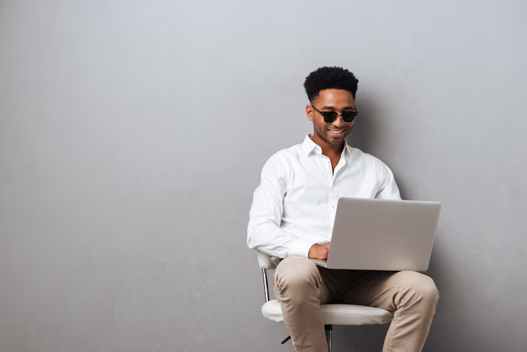 homem negro sentado vestido camisa branca e calça pege trabalha com um computador descansando nas pernas.