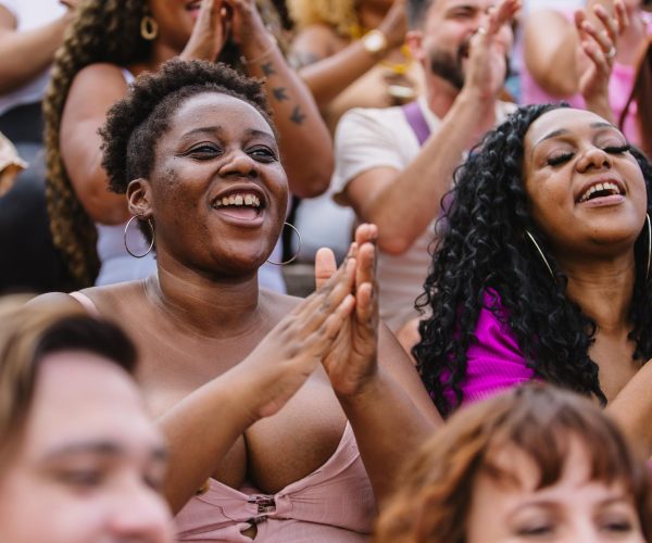 Uma multidão diversa participa de um evento ao ar livre, onde duas mulheres negras, sorridentes e demonstrando entusiasmo, aplaudem com energia. O ambiente é alegre e acolhedor, refletindo o acesso e a inclusão de mulheres negras e indígenas em espaços de destaque e celebração. A imagem ressalta o empoderamento feminino e a diversidade, com foco na representatividade de mulheres de diferentes origens étnicas em eventos públicos e culturais