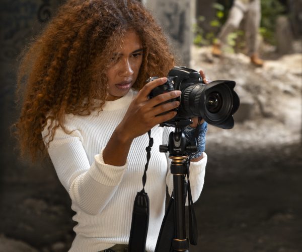 Uma jovem profissional negra do setor audiovisual ajusta sua câmera em um tripé enquanto se concentra na captura da imagem. Com cabelos volumosos e vestindo roupas casuais, ela está em um ambiente urbano e rústico, onde a arte e a técnica do audiovisual se encontram para contar histórias autênticas. Ao fundo, um homem desfocado caminha, acrescentando profundidade à cena. A imagem destaca a representatividade no audiovisual, evidenciando como a diversidade de olhares pode transformar narrativas e carreiras digitais.