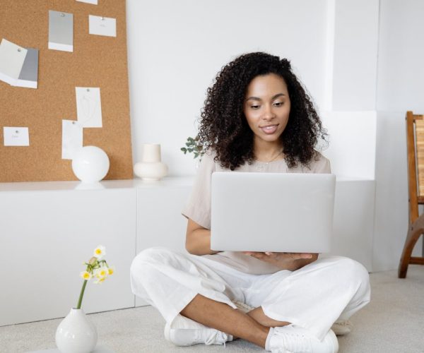 Cena de um ambiente minimalista, onde uma mulher está sentada no chão usando um notebook, representando um momento de planejamento e criação. Ao fundo, um painel de cortiça com recortes e amostras sugere inspiração visual e organização de ideias. O espaço harmonioso, com tons neutros e elementos decorativos simples, reflete um ambiente propício para o desenvolvimento de estratégias de branding criativas e focadas.