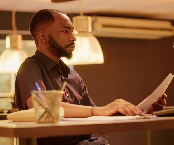 Homem negro de barba bem aparada, vestindo uma camisa social escura, está concentrado em sua mesa de trabalho. Com um olhar atento, ele segura um documento enquanto analisa informações no computador, em um ambiente de escritório iluminado por luminárias modernas. O espaço organizado, com itens como um caderno, lápis e pequenos vasos de plantas, transmite uma atmosfera profissional e produtiva, refletindo um momento de foco e estratégia. A cena representa o conceito de planejamento de carreira, destacando a importância da análise e organização para o crescimento profissional