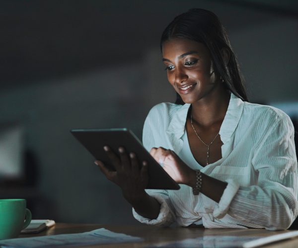Em um ambiente de trabalho com iluminação baixa, uma mulher negra com cabelos longos e lisos sorri enquanto utiliza um tablet. Ela veste uma camisa branca com amarração na frente e acessórios dourados. A luz da tela ilumina seu rosto, destacando sua expressão concentrada e confiante. Na mesa à sua frente, há uma xícara verde, documentos e um caderno. A cena transmite a força da comunicação quilombola no mundo digital, refletindo como as redes quilombolas utilizam a tecnologia para fortalecer narrativas negras, inovar e manter viva a tradição da resistência.