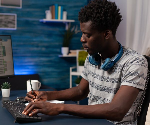 Homem jovem preto sentado à mesa, usando fones de ouvido e fazendo anotações em frente ao computador, simbolizando a descentralização da inovação.