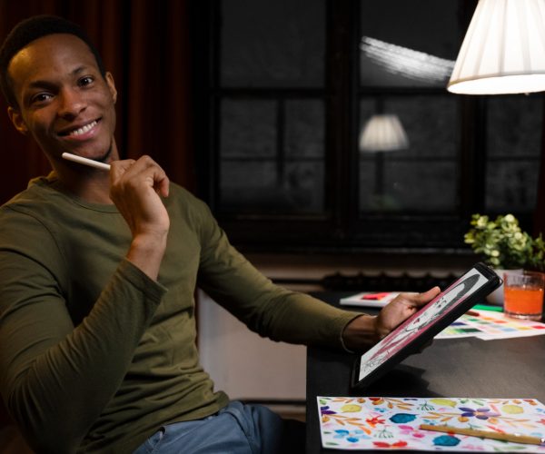 A imagem ilustra a economia criativa em ação, com um jovem artista digital sorridente segurando um tablet e uma caneta digital. Sentado em uma mesa com pinturas e ilustrações vibrantes ao redor, ele transmite a fusão entre arte tradicional e tecnologia. O ambiente aconchegante, iluminado por uma luminária pendente, ressalta a inspiração e a inovação como motores da criatividade. A cena representa o potencial econômico do setor criativo, onde talento e ferramentas digitais se unem para gerar valor e oportunidades.