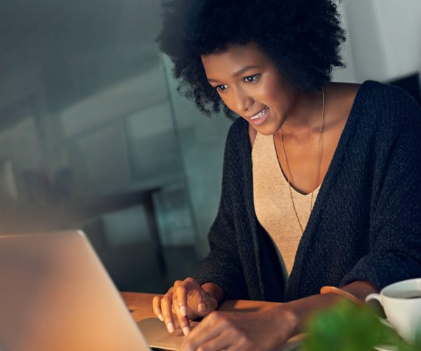 Uma jovem profissional negra, concentrada e sorridente, trabalha em seu laptop em um ambiente de escritório, simbolizando o empoderamento feminino na tecnologia. Papéis, caneta e uma xícara sobre a mesa reforçam seu foco e criatividade, destacando a importância da presença de mulheres em projetos de programação e inovação