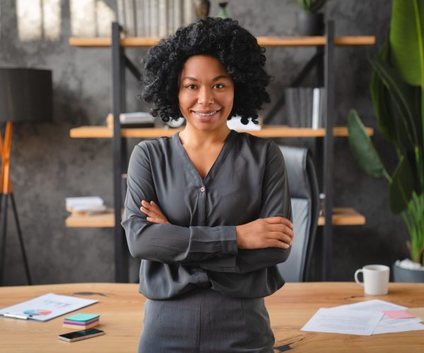 Uma mulher negra, com expressão confiante e postura ereta, está de braços cruzados em frente a uma mesa de escritório, simbolizando a liderança e a **gestão de equipes** em um ambiente corporativo. Ela veste uma camisa social cinza e sorri levemente, transmitindo segurança e profissionalismo. Ao fundo, vê-se uma estante com objetos decorativos e plantas, reforçando a atmosfera moderna e organizada do espaço de trabalho.
