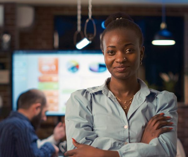 Mulher preta de expressão confiante, vestindo uma camisa azul-clara e com os braços cruzados, se destaca em um ambiente corporativo moderno. Ao fundo, uma tela exibe gráficos e dados de desempenho, enquanto colegas trabalham. A imagem simboliza a atuação de uma líder de marketing generalista, demonstrando estratégia e protagonismo no setor.