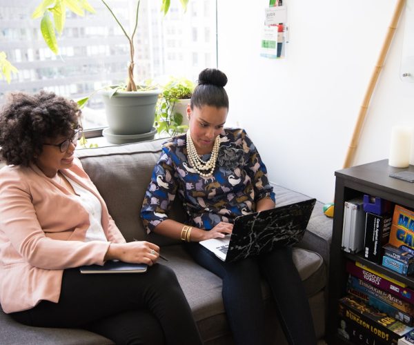 Duas mulheres sentadas em um sofá de escritório discutem ideias enquanto olham para um laptop. Uma delas usa um blazer rosa e segura um caderno, enquanto a outra usa uma blusa estampada e um colar de pérolas. Ao fundo, há plantas, uma prateleira com jogos e livros, e uma lousa branca com anotações. A cena reflete um ambiente colaborativo e acolhedor, simbolizando como marcas com propósito podem criar espaços que incentivam criatividade, inclusão e impacto positivo.