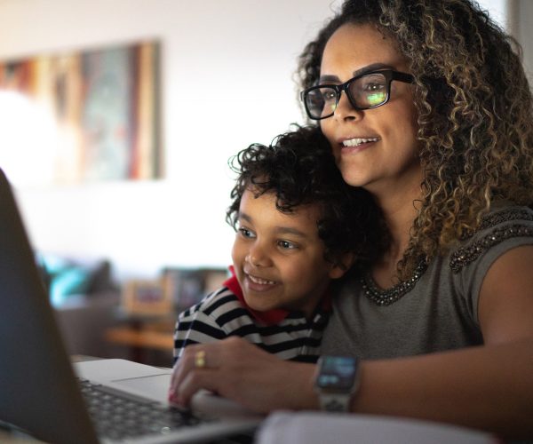 A imagem mostra uma mulher, com uma criança no colo, na frente de um notebook. São pessoas pretas e demonstram alegria. A imagem quer representar como a mulher lida com a maternidade e carreira.