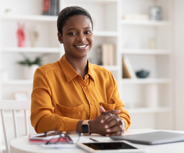 A imagem traz uma mulher preta, de cabelos curtos, vestindo uma blisa social amarela, sentada em uma mesa branca onde etão um óculos de grau, um notebook e um tablet. Essa imagem busca representar uma mulher confiante após fazer a migração de carreira