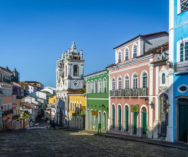 Rua do Pelourinho, em Salvador, Bahia, com seu calçamento de pedra e casarões coloridos que refletem a riqueza da arquitetura colonial brasileira. Ao fundo, a igreja barroca de fachada branca se destaca, representando a mistura de culturas e a ancestralidade presente nas influências africanas, europeias e indígenas que moldaram a história e o patrimônio cultural da região. O céu azul complementa a atmosfera vibrante e histórica do local.