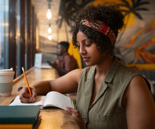 Mulher preta, com cabelo cacheado, usando uma faixa estampada, sentada em um ambiente de cafeteria, escrevendo em seu caderno com foco. Ao lado dela, há uma xícara de café reutilizável. A imagem reflete um momento de planejamento semanal, em que ela organiza suas atividades com concentração e determinação, aproveitando o ambiente acolhedor para se preparar para os desafios da semana