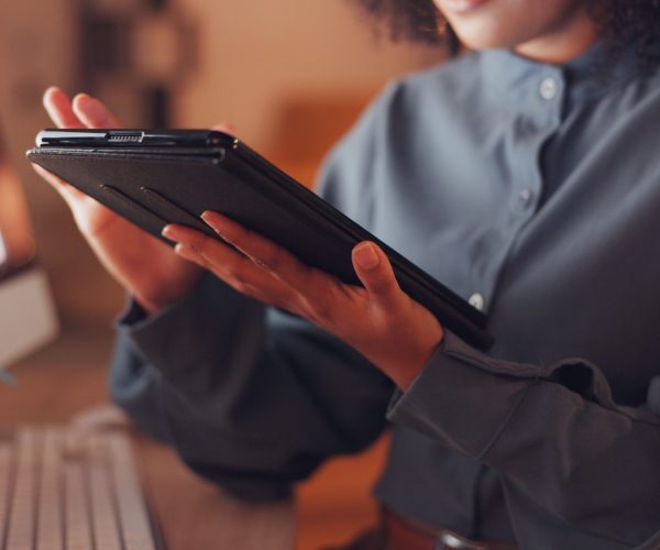 Uma mulher preta, vestida com uma camisa cinza, segura um tablet enquanto trabalha em um escritório como social media. Ela está focada em criar e analisar estratégias para redes sociais, demonstrando concentração em suas tarefas diárias.